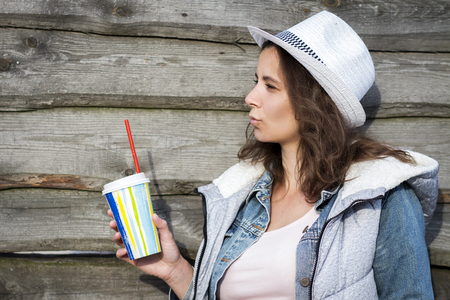Stylish young attractive girl in hat holds glass of cocktail drink on wooden wall backgroundの写真素材