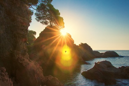 Scenic sunrise on sea coast in Spain, Costa Brava. Morning sea landscape in Lloret de Mar. Sun shines behind rock in mediterranean bayの写真素材