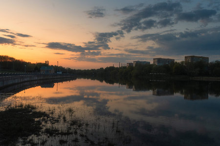 A peaceful evening on a Minsk lakeside. The colorful sunset sky, painted in hues of orange and purple, is mirrored perfectly in the calm water, with the illuminated city skyline creating a modern silhouette.の写真素材