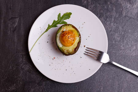 Cooked avocado with egg on a white plate on a dark background. A fork is on the plateの写真素材