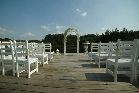 Venue of the wedding ceremony: a white arch decorated with flowers, white chairs for guests on the background of the lake and forestの写真素材