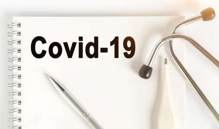 On the table are a stethoscope, a thermometer, a pen and a notebook with the inscription -Covid-19....