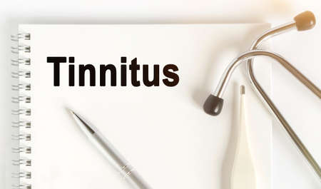 On the table are a stethoscope, a thermometer, a pen and a notebook with the inscription -Tinnitus. The concept of medicine.の写真素材