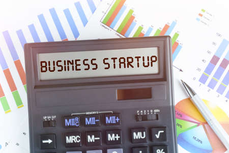 The concept of business and finance. On the table are financial charts and a calculator, on the electronic board of which is written the text - BUSINESS STARTUPの写真素材