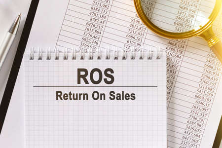 On the table are financial reports, a pen, a magnifying glass and a notebook with the inscription - ROS. Return On Sales. Business and finance concept.の写真素材