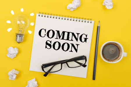 Business and finance concept. On the yellow table are: a light bulb - an idea, a cup of coffee, pieces of paper, glasses and a notebook with the inscription - COMING SOONの写真素材
