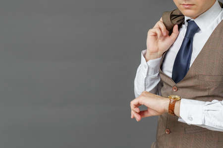 Portrait of a businessman who looks at his watch and holds a jacket over his shoulder. The face is not visible. Gray background. Business and finance conceptの写真素材