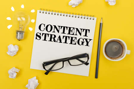 Business and finance concept. On the yellow table are: a light bulb - an idea, a cup of coffee, pieces of paper, glasses and a notebook with the inscription - CONTENT STRATEGYの写真素材