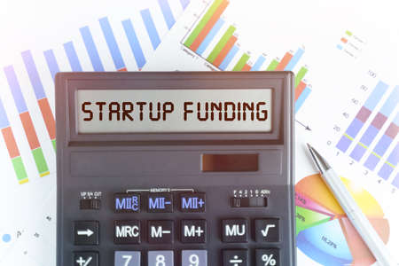 The concept of business and finance. On the table are financial charts and a calculator, on the electronic board of which is written the text - STARTUP FUNDINGの写真素材