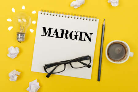 Business and finance concept. On the yellow table are: a light bulb - an idea, a cup of coffee, pieces of paper, glasses and a notebook with the inscription - MARGINの写真素材