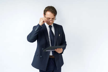 Portrait of a businessman in a blue suit who is holding an electronic tablet in his hands and adjusting his glasses. White background. Business and finance conceptの写真素材