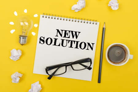 Business and finance concept. On the yellow table are: a light bulb - an idea, a cup of coffee, pieces of paper, glasses and a notebook with the inscription - NEW SOLUTIONの写真素材