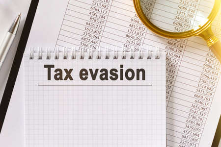On the table are financial reports, a pen, a magnifying glass and a notebook with the inscription - Tax evasion. Business and finance concept.の写真素材