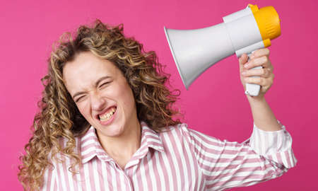A young woman holds a screaming megaphone in her hands, which is aimed at her face. Isolated pink background. People sincere emotions lifestyle concept.の写真素材