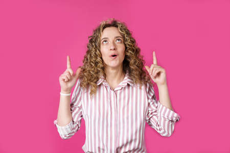 Young beautiful woman, amazed and surprised, looking up and pointing with fingers and raised hands. Isolated on pink background.の写真素材