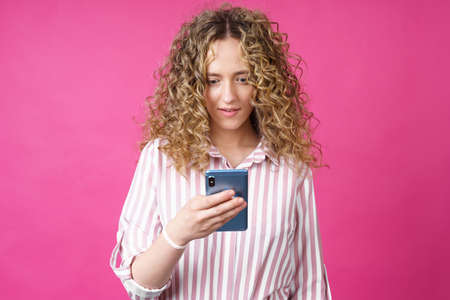 A fashionable woman in a striped shirt, holds a mobile phone, gasps in surprise, reads amazing news. Isolated on pink backgroundの写真素材