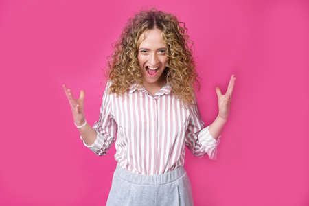 A curly-haired woman raises her palms in joy, glad to receive an amazing gift from someone, screams loudly, wearing a striped shirt. Isolated on a purple background.の写真素材