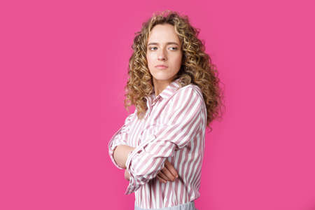 A side shot of a beautiful upset woman with curly hair holding her arms folded. Isolated on pink background.の写真素材
