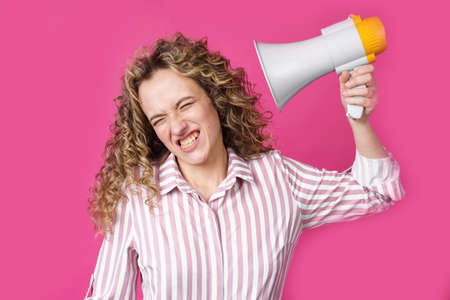 A young woman holds a screaming megaphone in her hands, which is aimed at her face. Isolated pink background. People sincere emotions lifestyle concept.の写真素材