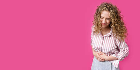 The young woman closed her eyes in pain, holding her stomach. Isolated on pink background