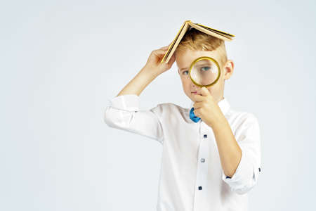 The schoolboy put the book on his head and looks through a magnifying glass. Isolated background. Education conceptの写真素材