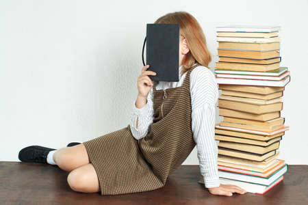 Education concept. A teenage girl sits on a table near books, covering her face with a notebook.