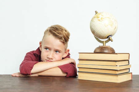 Education concept. A tired schoolboy sits at the table, put his head on his hands. On the table there are books, a globe and an alarm clock.の写真素材