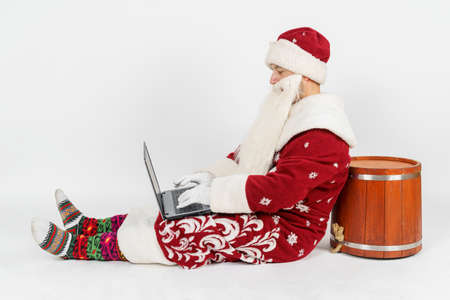 Christmas and New Years concept. Santa Claus is sitting on the floor and working at a laptop. Isolated background.の写真素材