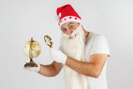 Christmas and New Years concept. Santa Claus is studying the globe with a magnifying glass. Isolated background.の写真素材