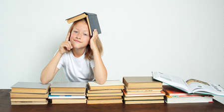 Education concept. The student sits at the table, with a textbook on her head. She raised her finger emotionally. Books are spread out nearbyの写真素材