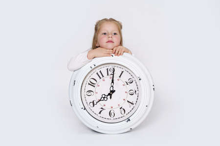 Children concept. The girl is sitting on the floor in front of a large white clock. Isolated over...