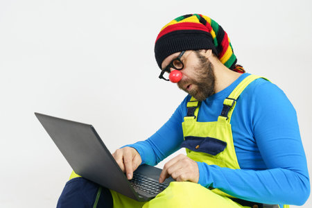 Celebration and communication concept. A clown in bright clothes sits on the floor and communicates via a laptop.の写真素材