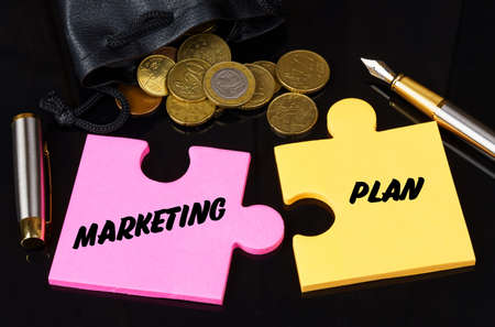 Business and finance concept. Coins are scattered on the black surface, there is a pen and puzzles on which it is written - MARKETING PLANの写真素材