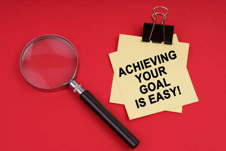 Business and Economics. On a red background, a magnifying glass and a sticker with the inscription - Achieving your goal is easyの写真素材