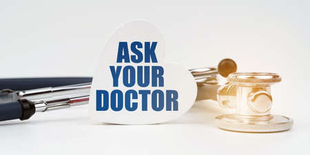 On the white surface there is a stethoscope and a heart plate with the inscription - ASK YOUR DOCTORの写真素材