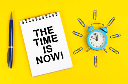business concept. On a yellow surface there is an alarm clock, a pen and a notepad with the inscription - The Time is Nowの写真素材