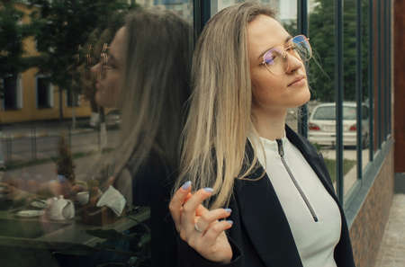 A beautiful woman stands leaning against the window of a building, touches her hair with her hands, looks into the distance. Portrait of a beautiful woman.の写真素材