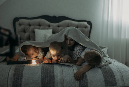 Mom in the evening with a flashlight reads a book to children covering them with a blanket. close-up.の写真素材
