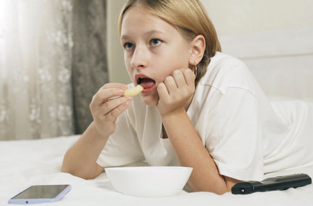 A teenage girl lies on a bed, eats corn sticks and looks into a smartphone and TV. close-up.