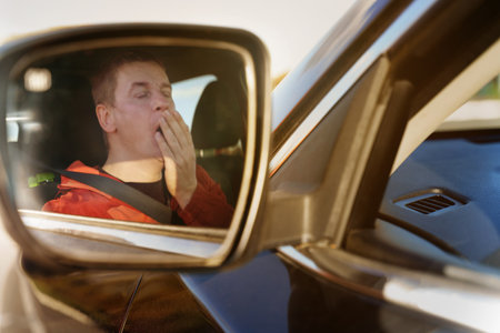 A tired driver yawns while covering his mouth with his hand while sitting at the wheel. View of the...