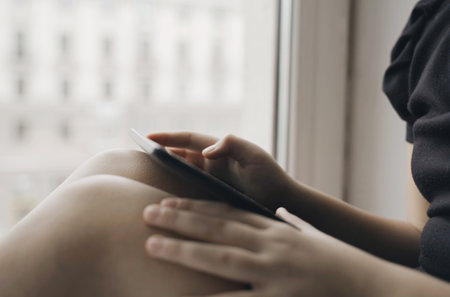 A teenage girl is sitting on a windowsill in her hands with a tablet, she is studying, watching a...