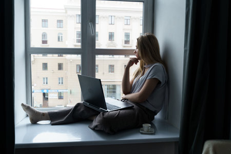 A young woman sits on a windowsill, works at a laptop, looks out the window. business and education...