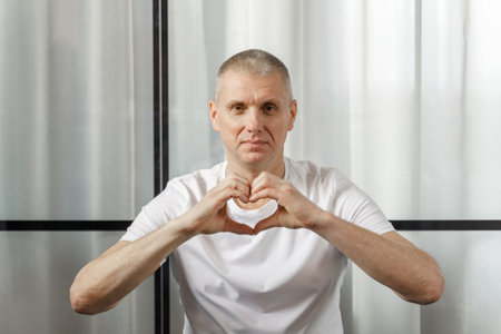 A middle-aged man in a white T-shirt smiles and shows a heart symbol.の写真素材