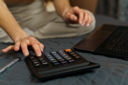 A young woman sits on the bed, uses a calculator and a laptop to work in the bedroom at home....