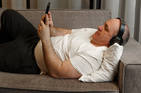 A middle-aged man lies on the couch, listens to music on headphones, looks at the phone.の写真素材