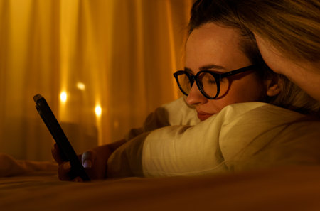 Young woman in glasses with smartphone lying in bed at home at night. close-up