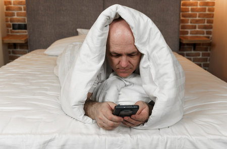 An adult man lies covered with a blanket and browses social networks, videos in a smartphone.の写真素材