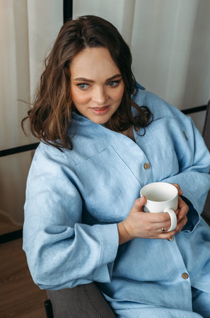 Portrait of a young woman in a light blue suit, who is sitting on a chair with a white cup in her hands. vertical frame.の写真素材