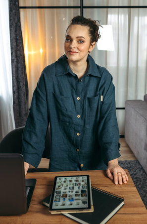 Portrait of a business woman standing near the desk. Notepad, tablet and laptop on the table....