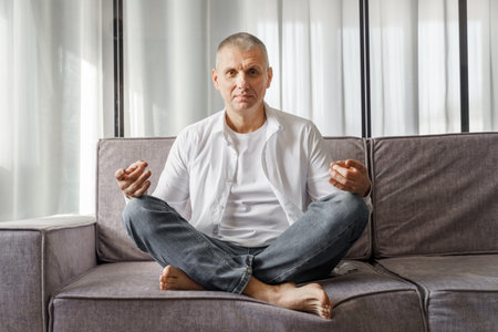 Portrait of a man who sits on a sofa in a room in a lotus position. The man is doing yoga.の写真素材